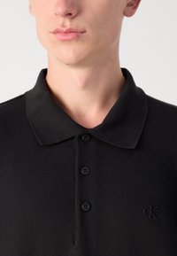 Črna polo majica iz pletenine, z ovratnikom, tremi gumbi in majhnim, vrezanim logotipom na levi strani prsnega koša.