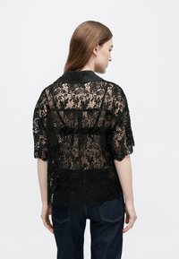 Sort bluse i blonder med korte ærmer og en satin krave. Stoffet har blomsterudskæringer og en halvgennemsigtig tekstur.