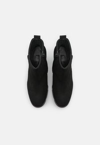 Para czarnych zamszowych botków za kostkę z bocznymi zamkami błyskawicznymi i pętlami do ciągnięcia, widok z góry na białym tle.