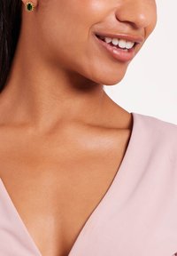 Oorbellen met een smaragd geslepen groene steen en gouden accenten, in een delicate ontwerp, tentoongesteld tegen een zachte roze stofachtergrond.