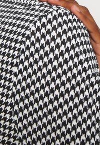 Ruka odpočívajíc�í na látce s černobílým vzorem houndstooth, ukazující podrobnou texturu a detailní záběr na prsty a palec.