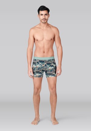 Jonge man met kort donker haar staat op blote voeten naar voren gericht, draagt groene boxershort met camouflagepatroon, op een effen grijze achtergrond.