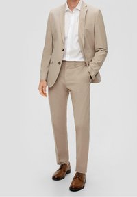 Bežový oblek se skládá z propracovaného saka a kalhot na míru, doplněných bílou košilí a hnědými šněrovacími botami. Hladký povrch látky.