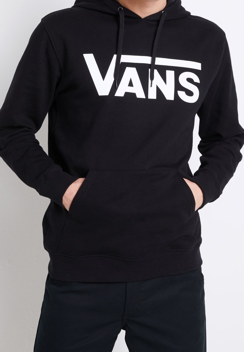 Personne portant un sweat à capuche Vans noir avec logo blanc, mains dans la poche avant, associé à un pantalon noir, fond clair uni.