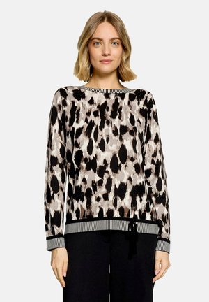 Femme portant un pull à manches longues à imprimé léopard avec des poignets et un ourlet rayés, associé à un pantalon noir, debout devant un fond uni.