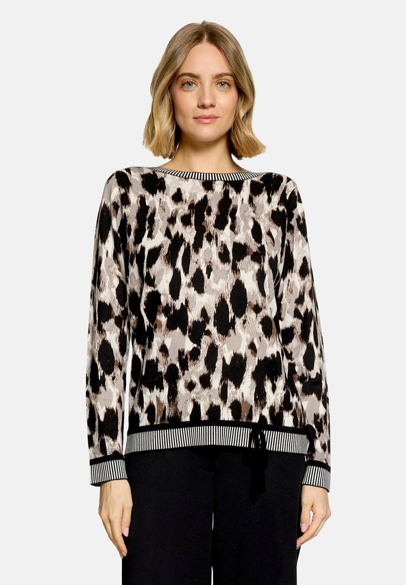 Femme portant un pull à manches longues à imprimé léopard avec des poignets et un ourlet rayés, associé à un pantalon noir, debout devant un fond uni.