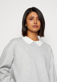 Sweatshirt gris clair avec un col blanc texturé, comportant des manches longues et une coupe décontractée. Tissu doux et détails de design simples.