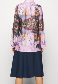 Hellrosa Bluse mit langen Ärmeln und floralem sowie fruchtigem Muster, ausgestattet mit einer halbtransparenten Textur. Kombiniert mit einem dunkelblauen, knielangen Rock.