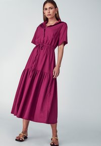 Maksi haljina u burgundi boji, izrađena od mekog materijala. Ima kratke rukave, kopčanje na dugmadi sprijeda, struk s naborima i slojevitu suknju.