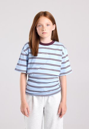 Fille aux cheveux rouges et raides portant un t-shirt à manches courtes rayé bleu clair et foncé et un pantalon gris clair, debout devant un fond uni.