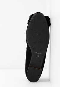 Chaussure plate noire avec semelle à motif montrant de petites empreintes et la taille 38, positionnée debout contre un mur blanc d'angle.