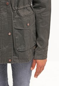 Gros plan d’une personne portant une veste vert foncé avec des boutons-pression en cuivre et une grande poche avant, associée à un jean bleu clair.