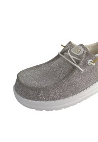 Glittering silver shoe with a rounded toe, white rubber sole, and decorative white laces. Branding label visible on the tongue.