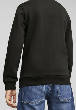 Personne portant un sweat-shirt noir uni et un jean en denim bleu, vue de dos sur un fond gris clair uni.