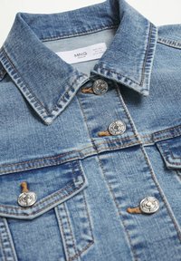 Veste en jean bleue avec boutons métalliques argentés, coutures marron, col et deux poches poitrine boutonnées, affichant l’étiquette de la marque MNG à l’intérieur.