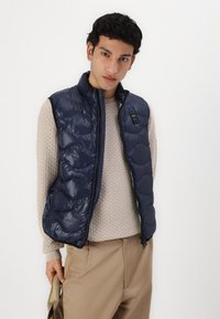 Navy blå puffvest med høy krage og glidelås. Teksturt beige genser under og beige bukser. Diskret merkevaredetalj synlig.