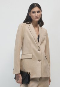 Donna che indossa una giacca beige su misura e pantaloni, mentre tiene una borsa a tracolla nera con una mano tatuata visibile su uno sfondo chiaro.