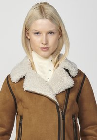Giacca finta suade marrone con un colletto di shearling, chiusura con zip e tasche laterali. Sotto è indossato un dolcevita crema a coste.
