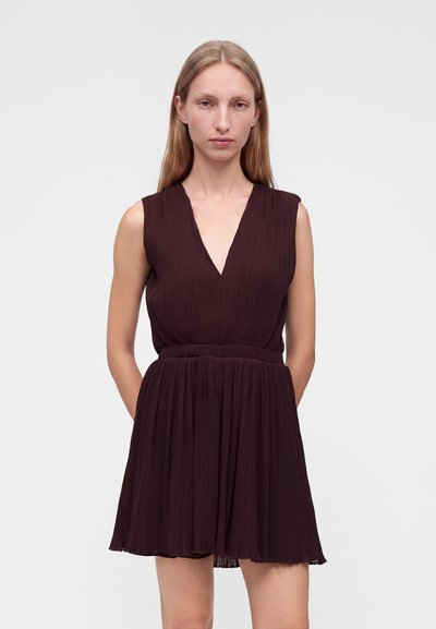Femme aux longs cheveux blonds portant une robe plissée marron foncé sans manches avec un profond décolleté en V, debout devant un fond clair uni.
