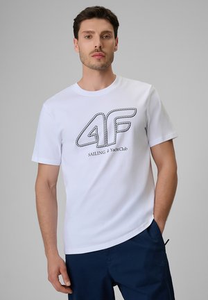 Mężczyzna w białej koszulce z logo "4F Sailing Yacht Club" i granatowych spodniach na jednolitym szarym tle.