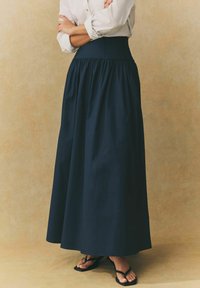 Jupe maxi bleu marine avec une taille cintrée et un tissu froncé. Texture lisse, design longueur cheville, associée à des tongs noires.