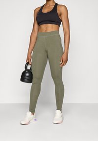 Top corto atlético negro, leggins verde oliva, tela texturizada, diseño ajustado. La modelo sostiene una kettlebell negra, vistiendo zapatillas blancas con detalles.
