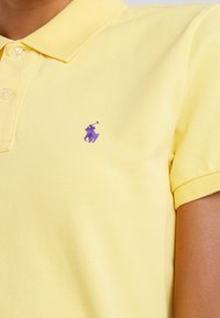 Žluté polo tričko vyrobené z texturované látky, s límečkem na dva knoflíky a malým fialovým vyšívaným logem polo hráče na hrudi.