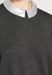 Zbliżenie osoby w ciemnoszarym swetrze z błyszczącym srebrnym wysmukłym kołnierzem na jednolitym tle.