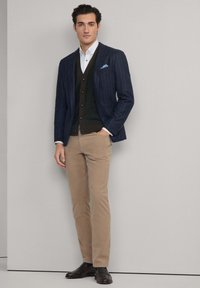 Marineblauwe blazer met streepjes, wit shirt, donkergroen vest, beige broek en bruine schoenen. De blazer heeft een lichtblauwe pochet.