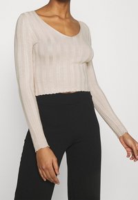 Top long à manches longues beige clair, côtelé, avec un col en V et une coupe courte. Présente une surface légèrement texturée et une silhouette ajustée. Associé à un pantalon noir.