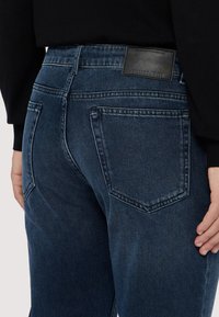 Jeans de mezclilla de color azul oscuro con un parche de cuero en la parte posterior. El diseño incluye dos bolsillos traseros con detalles de costura y un corte recto.