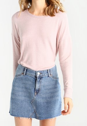 Žena v lehkém růžovém svetru s dlouhým rukávem zastrčeném do modré džínové mini sukně s opraným lemem.