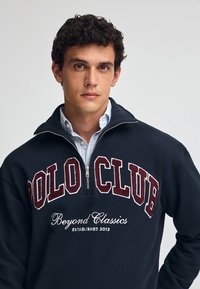 Pullovertröja i marinblått med design för kvartdragkedja. Har "POLO CLUB" broderat i bordeaux och "Beyond Classics" i vit text nedanför.