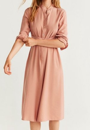 Femme portant une robe rose poudrée à manches longues, boutonnée, longueur genou, retroussant sa manche droite sur un fond uni.