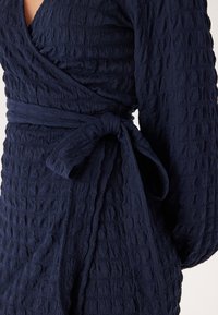 Marineblå texturerad omslagsklänning med långa ärmar, har en knut vid midjan och skrynklig tyg för extra detaljer och dimension.