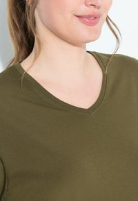 Olivově zelené tričko s výstřihem do V. Vyrobeno z hladké tkaniny, má krátké rukávy a přiléhavý střih. Jemný šev detail poblíž výstřihu.