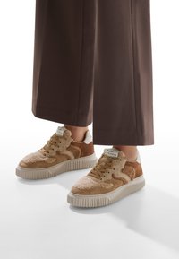 Baskets beiges et brunes avec une tige texturée ; en daim et matériau similaire à de la fourrure, une semelle blanche et des lacets bruns. Associées à un pantalon marron foncé.