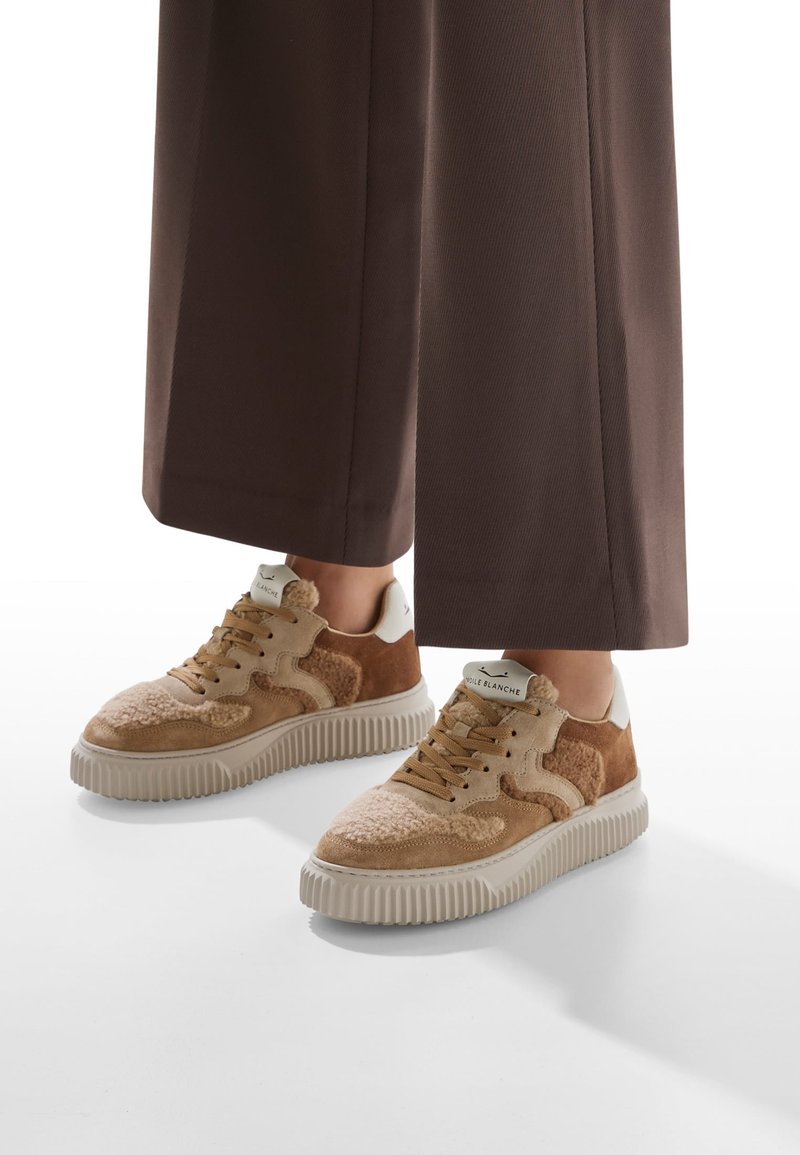 Baskets beiges et brunes avec une tige texturée ; en daim et matériau similaire à de la fourrure, une semelle blanche et des lacets bruns. Associées à un pantalon marron foncé.