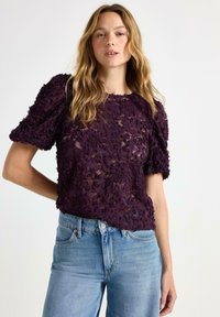 Lila blommig spetsblus med puffade kortärmar, halvgenomskinlig tyg, rund halsringning, kombinerad med ljusblå högmidjade jeans.