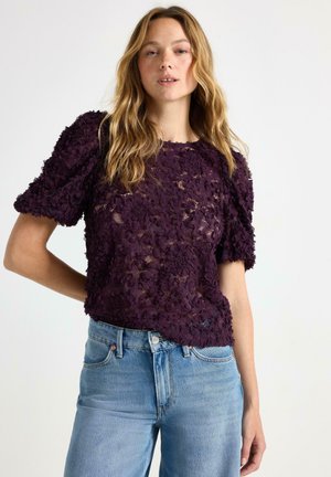 Lila blommig spetsblus med puffade kortärmar, halvgenomskinlig tyg, rund halsringning, kombinerad med ljusblå högmidjade jeans.