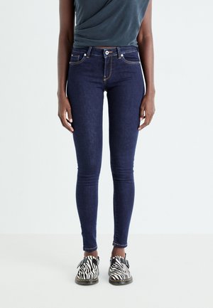 Person iført mørkeblå skinny jeans, en mørk indstøbt top og sort-hvide zebra-print sko, stående mod en ensfarvet lys baggrund.