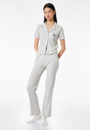 Jonge vrouw staand met handen achter haar rug, gekleed in een lichtgrijs kort mouw blouse met knopen en bijpassende broek met witte sokken.