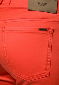 Jeans w kolorze koralu o gładkiej teksturze. Posiadają tylnią kieszeń oraz subtelny znak marki. Szwy są widoczne w kontrastowym kolorze.