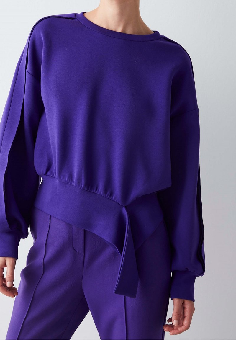 Personne portant un haut à manches longues violet vif avec de larges poignets et un pantalon assorti, debout devant un fond gris clair uni.