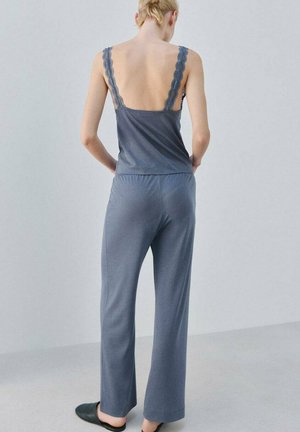 Femeie blondă purtând un maiou gri cu dantelă și pantaloni largi asortați, stând desculță pe un podea simplă de culoare gri deschis.