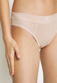 Bañador de bikini de color rosa claro con una cinturilla ancha que presenta un logo de adidas en blanco. El diseño es sin costuras y ajustado.