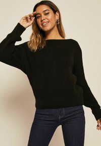 Pull en maille noir avec un large col bateau, texture côtelée et manches oversize, associé à un jean en denim foncé.