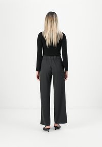 Una donna con lunghi capelli biondi si trova di fronte a una parete bianca, indossando una maglia nera a maniche lunghe, pantaloni scuri larghi e scarpe con tacco nere.