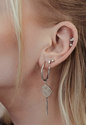 Nahaufnahme eines Ohrs mit vier silbernen Ohrringen: ein großer Creolenring mit einem kunstvollen Anhänger, zwei kleine Ohrstecker und ein hängender, blattförmiger Stecker am Knorpel.