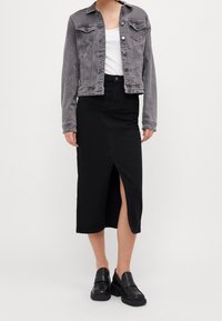 Giacca in denim grigia con chiusura a bottoni e due tasche sul petto, indossata sopra una maglietta bianca. Gonna nera con spacco frontale, abbinata a mocassini neri.